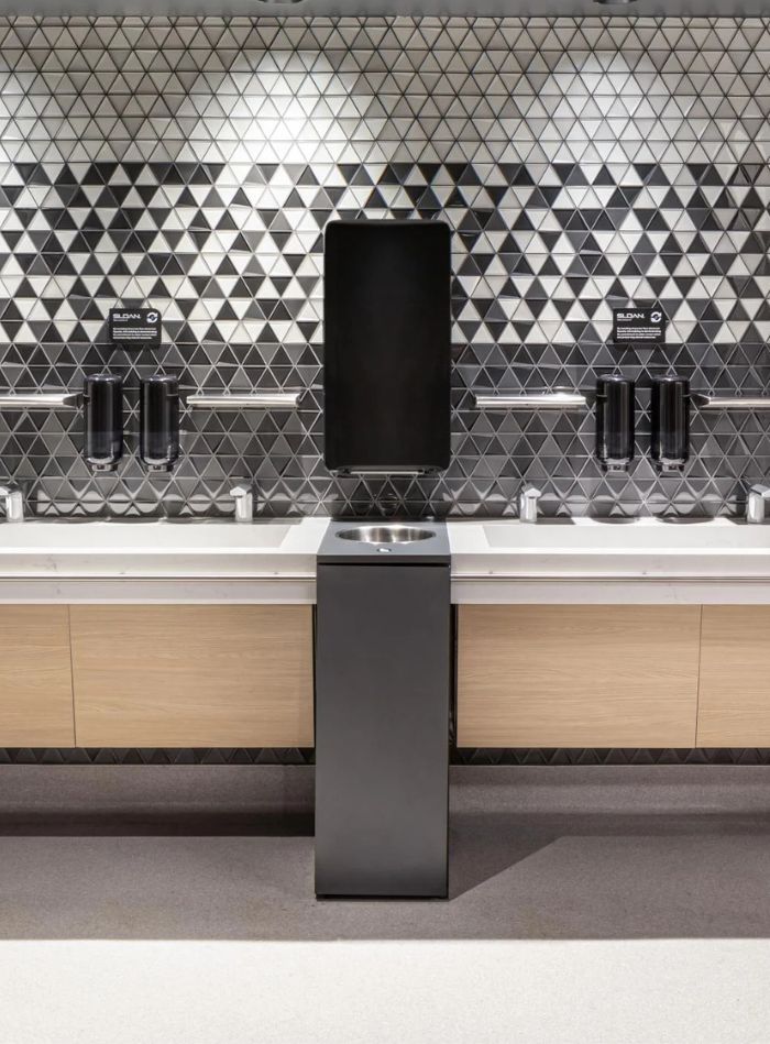 Modern sink area with light wood cabinetry and a black-and-gray triangular tile wall, featuring automatic dispensers and an integrated hand dryer.