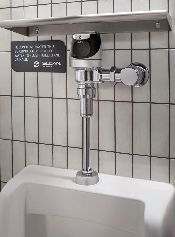 Automatic flushometer installed above a urinal, with a sign indicating the building uses recycled water to conserve resources.