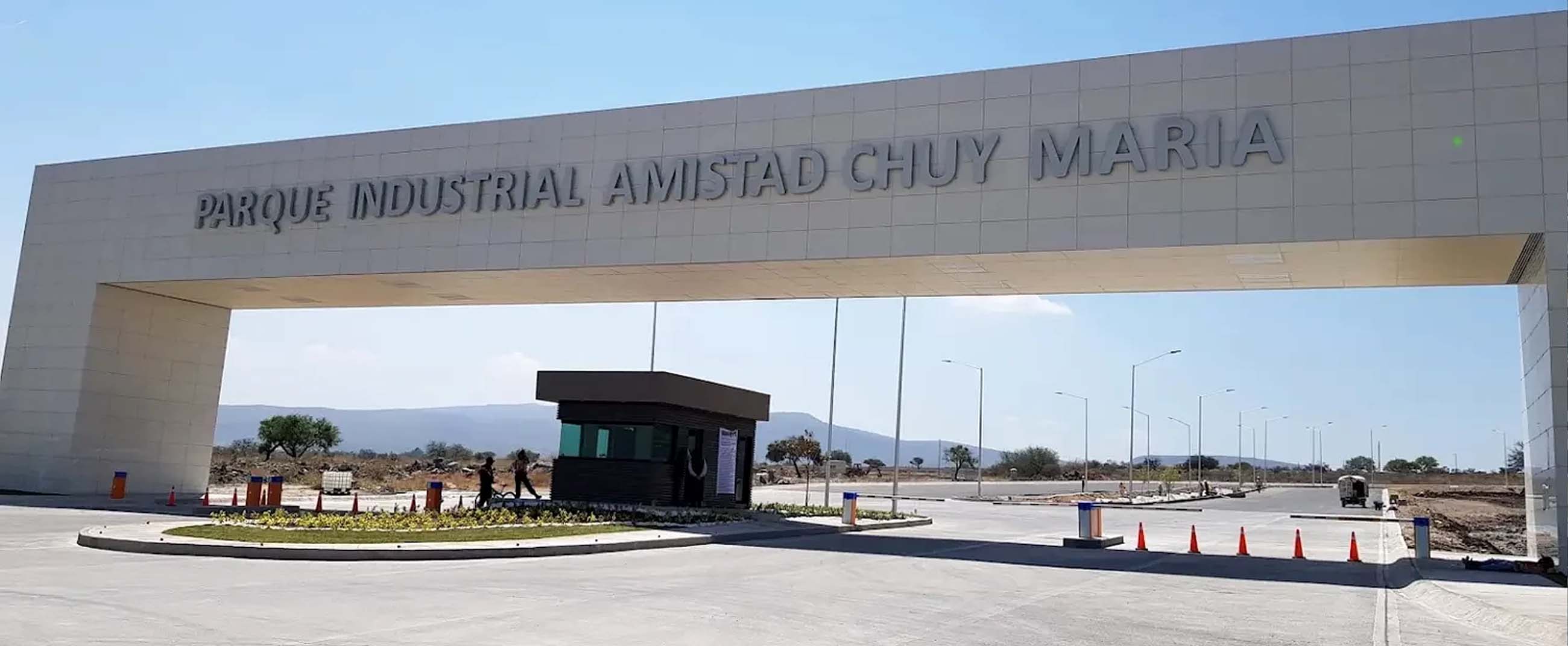 the main entrance of the Amistad Chuy María Industrial Park, a modern structure with a large light-colored gateway. At the center stands a security booth surrounded by wide, well-paved roads, with a semi-arid landscape visible in the background.