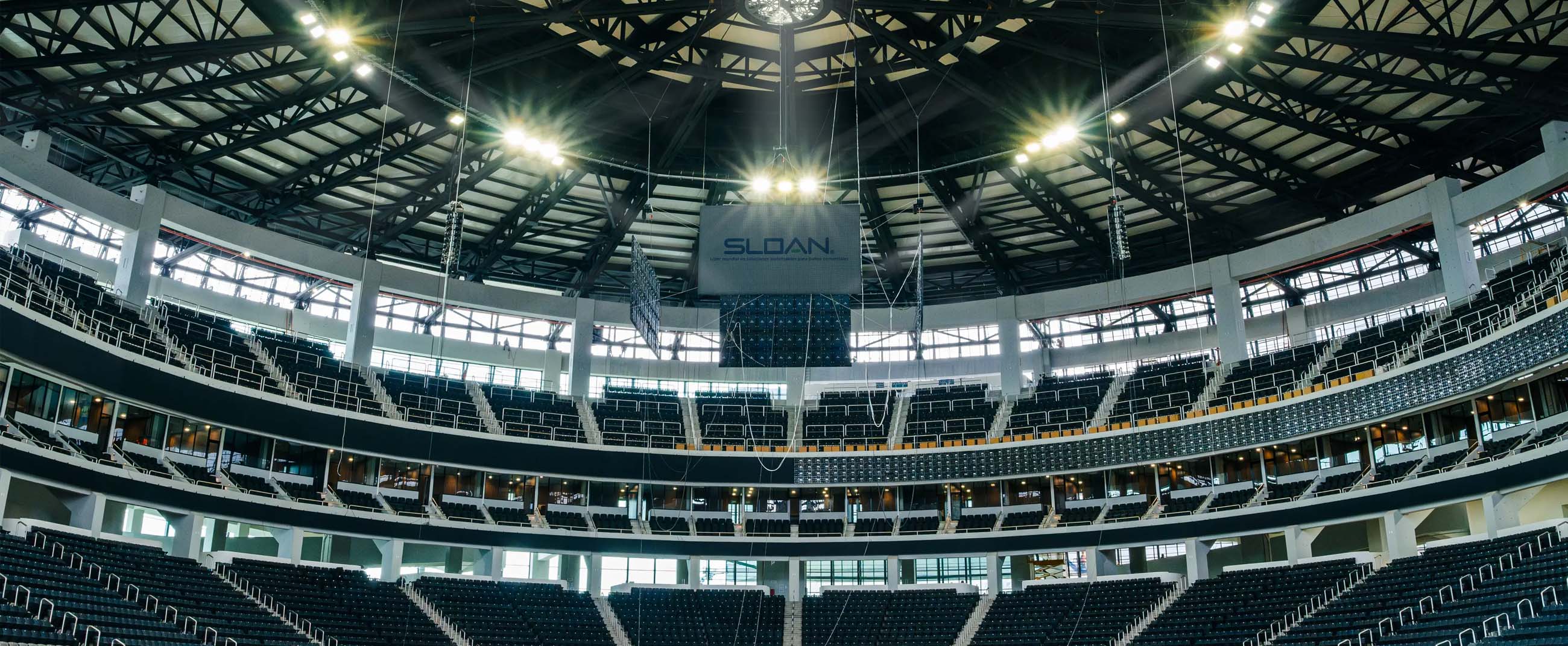 the interior of a modern sports arena with spacious seating and a steel-structured roof. A central screen bearing the Sloan logo hangs above, while the overhead lighting emphasizes the scale and architectural design of the venue.