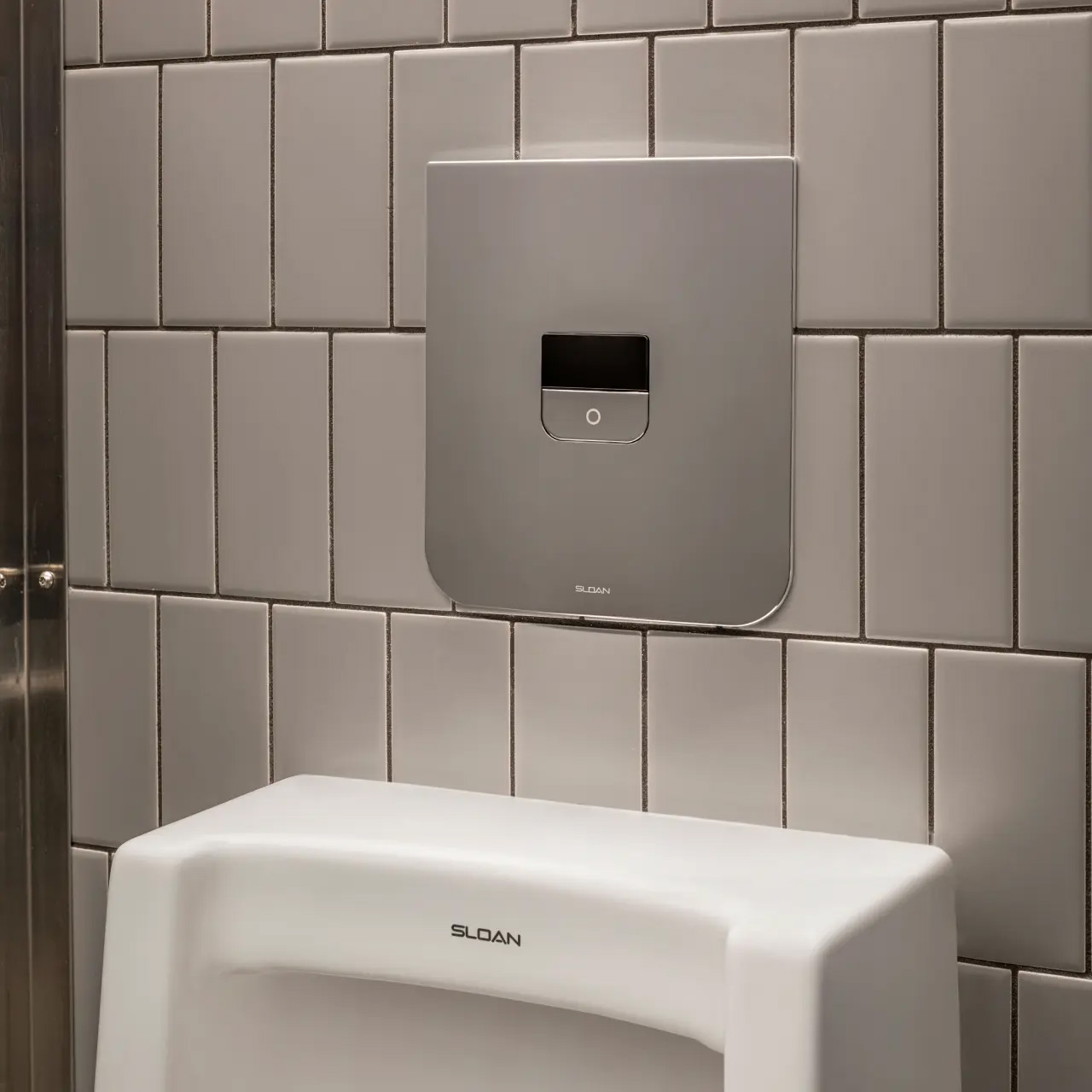 stainless-steel flush valve mounted on a wall of rectangular gray tiles. Below it, the upper portion of a white porcelain urinal with the Sloan logo is visible.