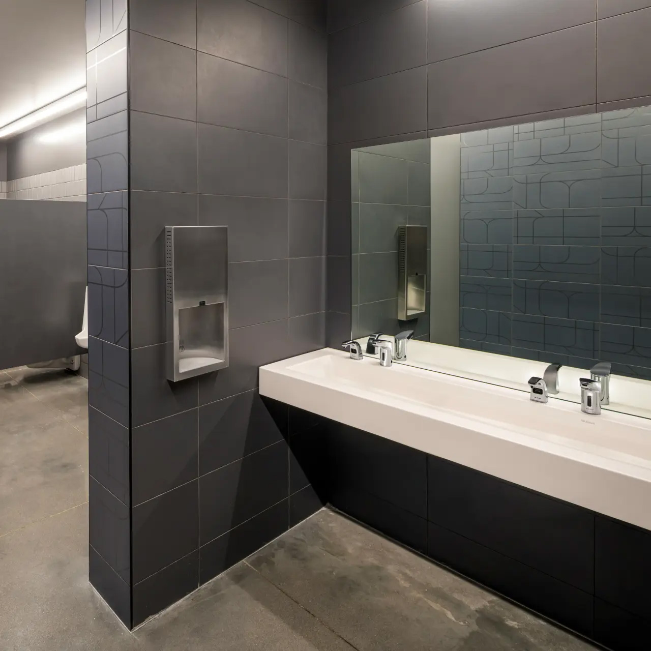 modern commercial restroom with a minimalist design, featuring matte gray tiled walls and a polished concrete floor. A continuous white sink with two chrome faucets sits beneath a large wall-mounted mirror that visually expands the space. To the side, a built-in stainless steel dispenser provides a clean, integrated look, while a partial urinal is visible in the background. Linear white lighting enhances the sense of cleanliness and highlights the space’s functional, contemporary aesthetic.