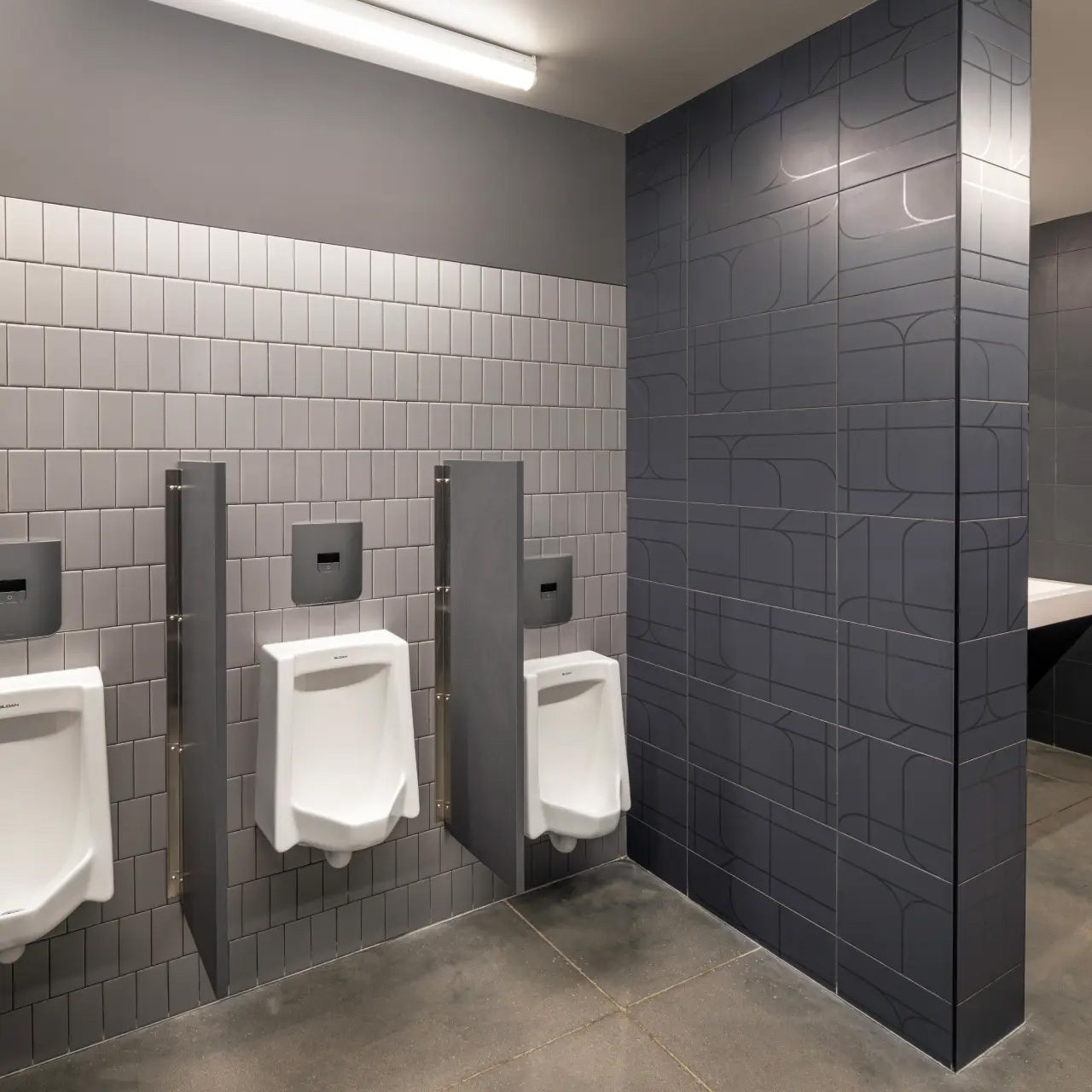 row of wall-mounted white urinals separated by gray dividers, set against a light gray tiled wall. Each unit features a built-in automatic flush valve, complemented by a polished concrete floor and a darker side wall with a subtle geometric pattern.