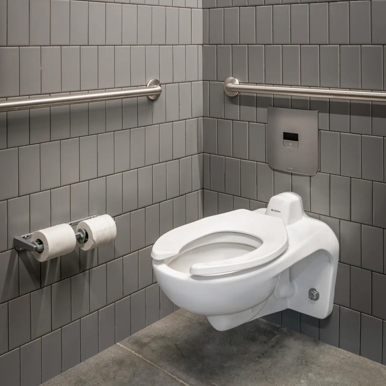 wall-mounted commercial toilet installed against gray rectangular tiled walls and a polished concrete floor. Stainless steel grab bars, a dual toilet paper holder, and a built-in sensor flush valve complete the setup. The overall design emphasizes functionality, accessibility, and hygiene, featuring modern lines and durable materials suited for public restrooms.
