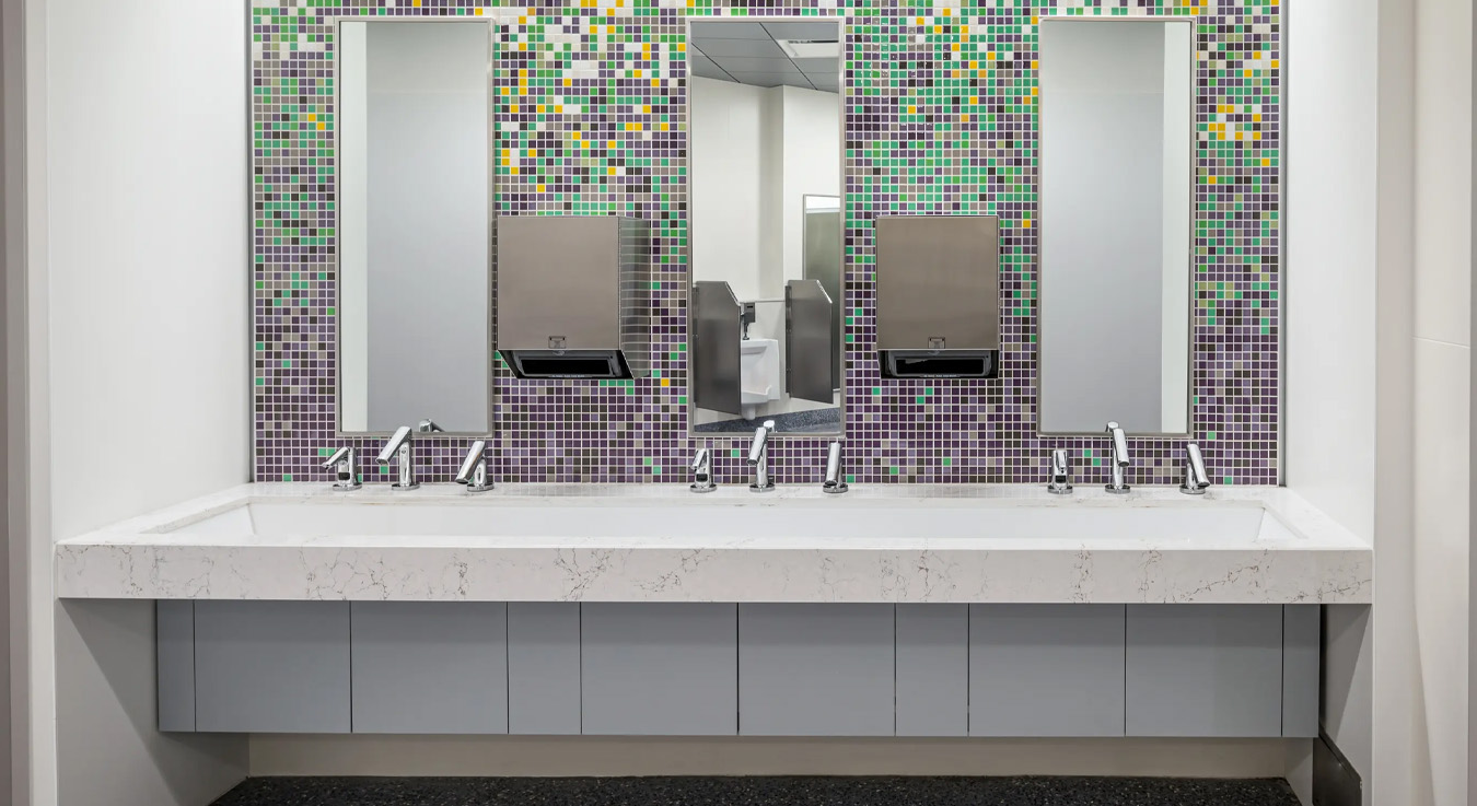 a modern washbasin area with a clean and colorful design. The back wall features a mosaic of small tiles in shades of green, purple, white, and yellow, adding an artistic touch to the space. The white marble countertop holds multiple sinks with stainless steel automatic faucets.
