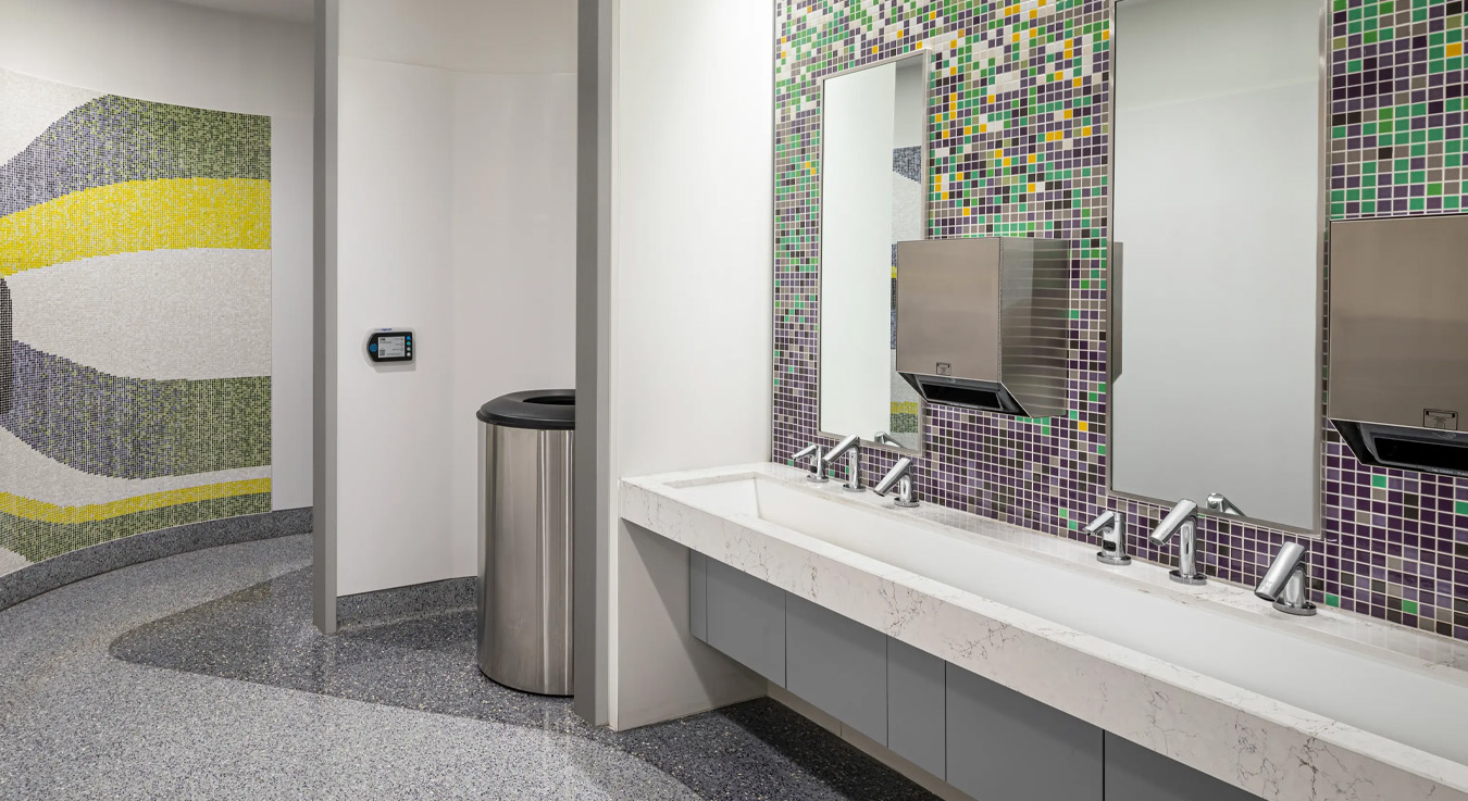 a modern public restroom with an artistic and clean design. In the foreground, white countertops with automatic faucets and stainless steel hand dryers can be seen. The back wall features colorful mosaic tiles in shades of green, purple, and yellow, adding a vibrant touch. In the background, a mural with flowing lines and a stylized human figure enhances the contemporary and functional atmosphere of the space.