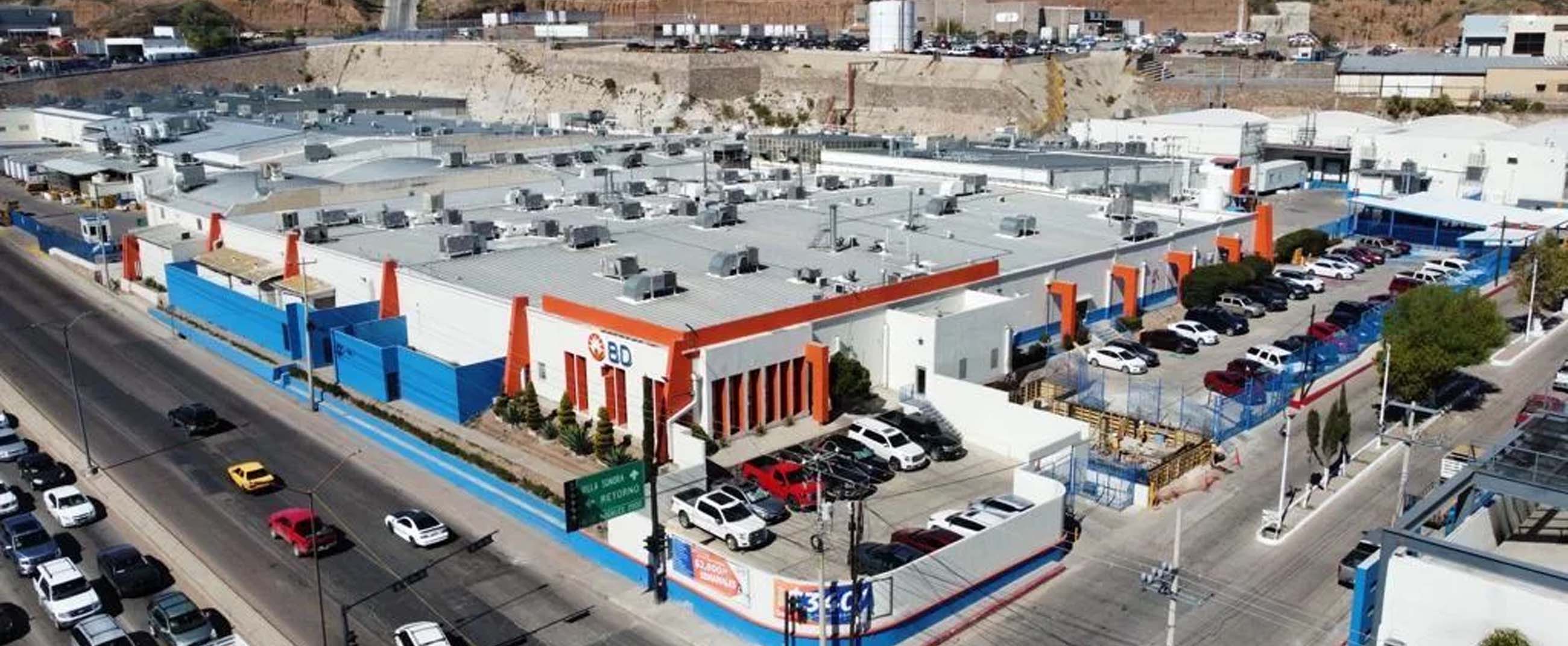 an aerial view of a modern industrial complex featuring large metal-roofed warehouses and an exterior design in white, blue, and orange tones. In the foreground, there are well-organized parking areas and a main avenue with light traffic, while in the background, urbanized hills and supporting structures complete the setting.