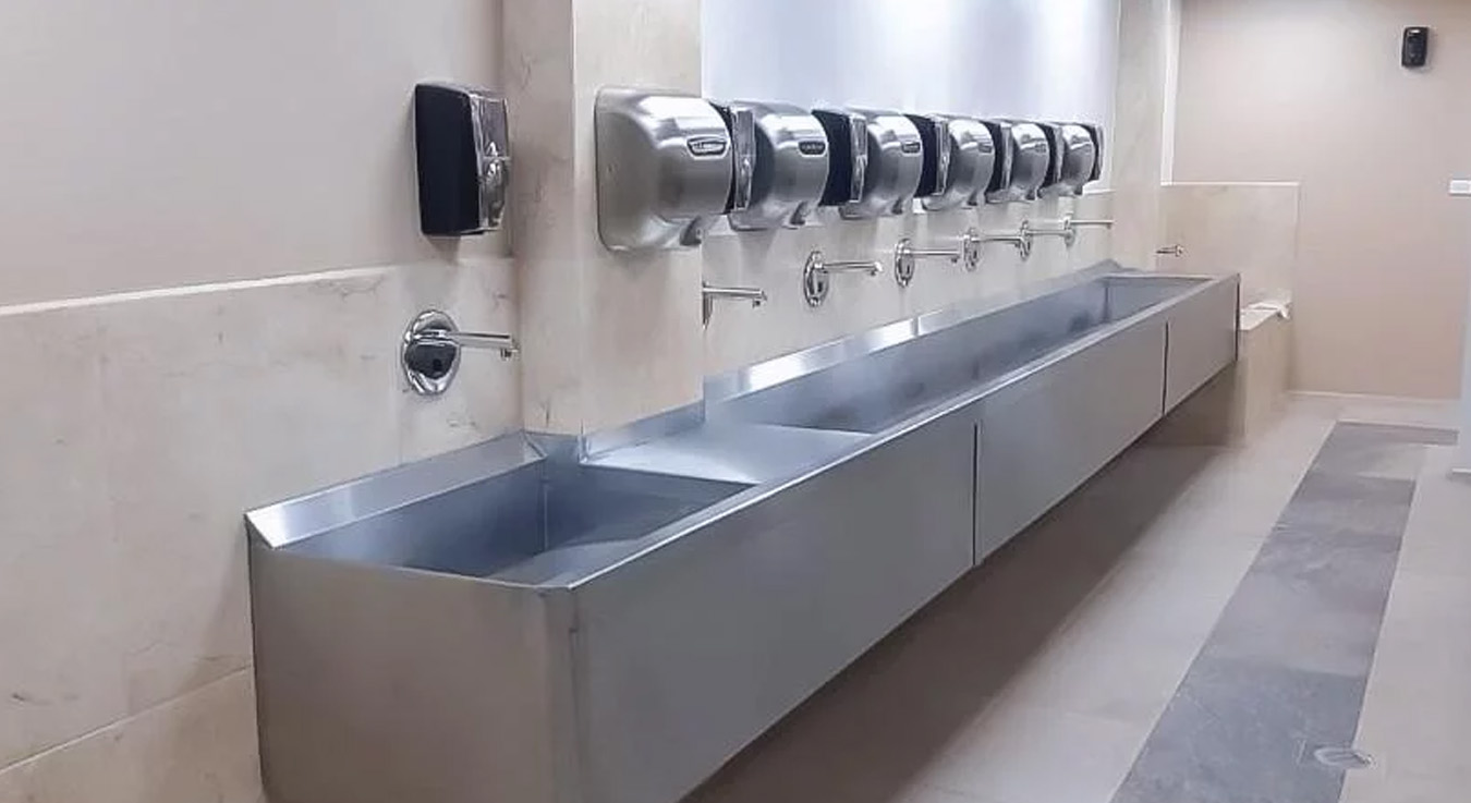 a modern stainless steel handwashing station equipped with multiple automatic soap dispensers, faucets, and integrated dryers. Its linear design and durable materials reflect a functional, hygienic, and long-lasting setup, ideal for high-traffic facilities.