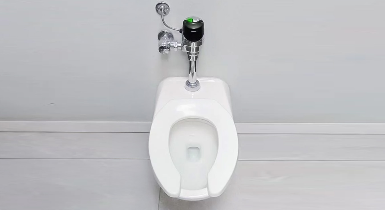 inodoro de uso público con asiento abierto al frente, equipado con un fluxómetro automático cromado de la marca Sloan. El diseño busca ofrecer higiene y eficiencia en el uso del agua, dentro de un entorno limpio y de acabados claros.