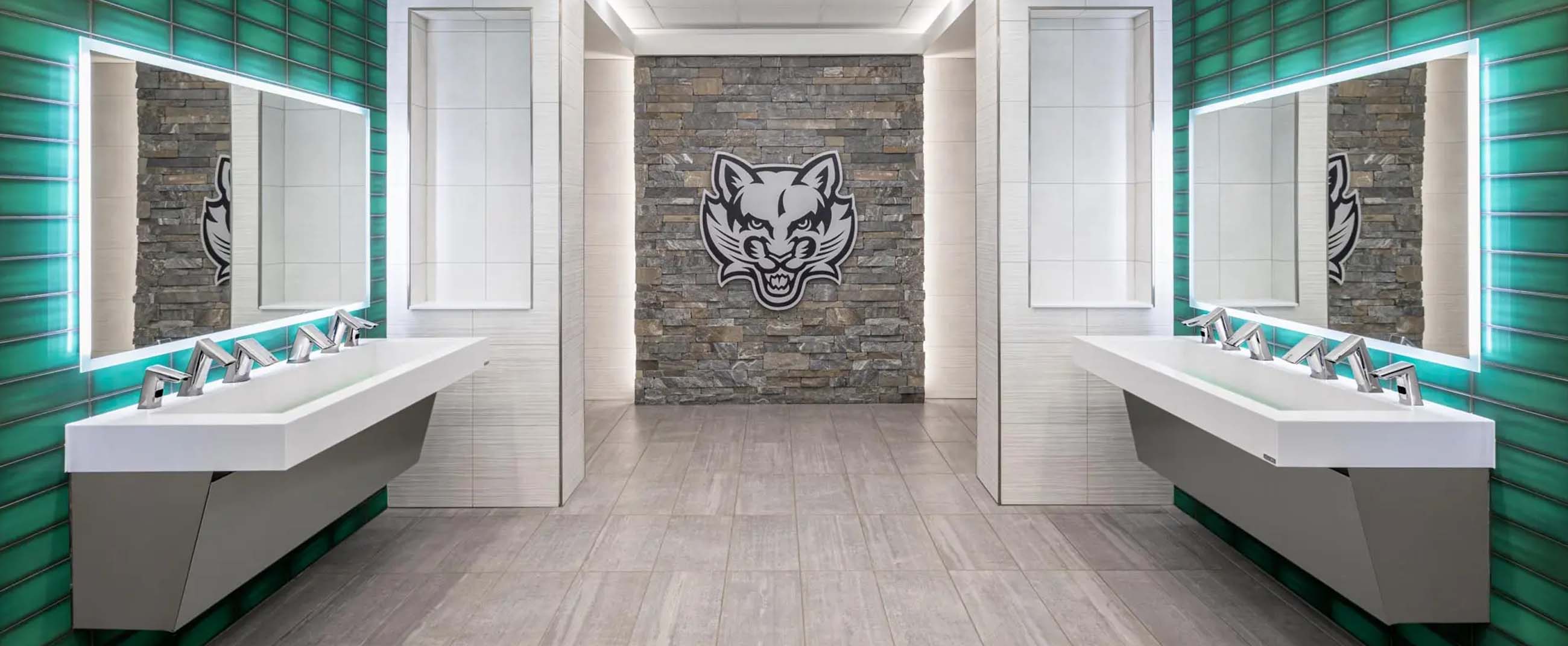 contemporary high-design restroom featuring two opposing double sinks, each with chrome automatic faucets and backlit mirrors. The side walls are clad in glossy green tiles that contrast with the light gray floor and central stone walls. At the back, a stone accent wall with a feline logo adds an institutional and athletic touch. Linear ceiling lighting enhances the symmetry, creating a modern, clean, and visually striking environment.