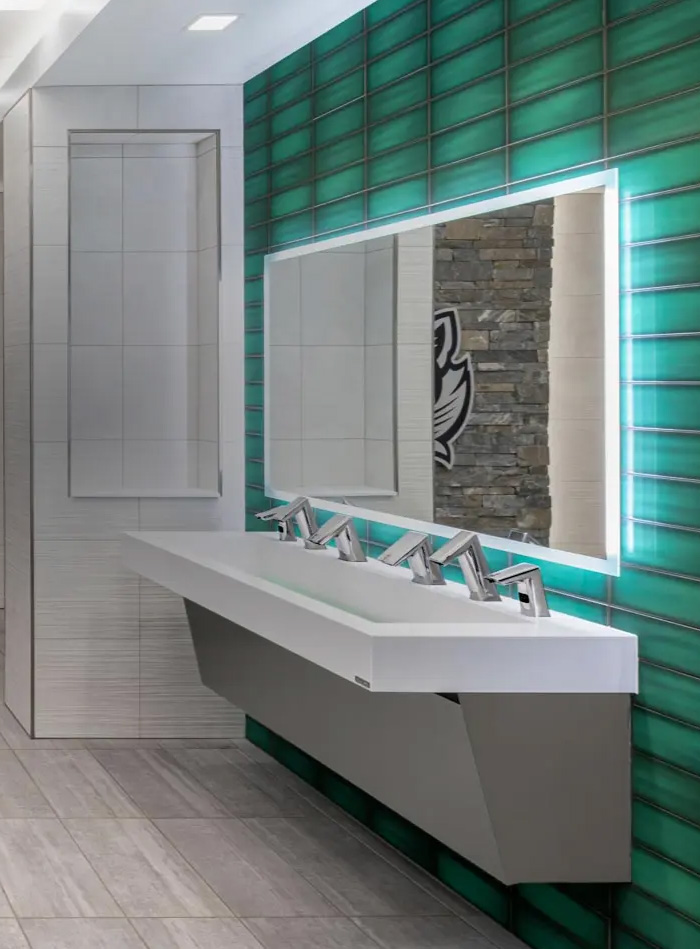 multi-station white solid-surface sink with Sloan chrome automatic faucets, set against a wall clad in glossy green tiles. A large mirror with integrated LED lighting enhances brightness and emphasizes the modern aesthetic of the space.