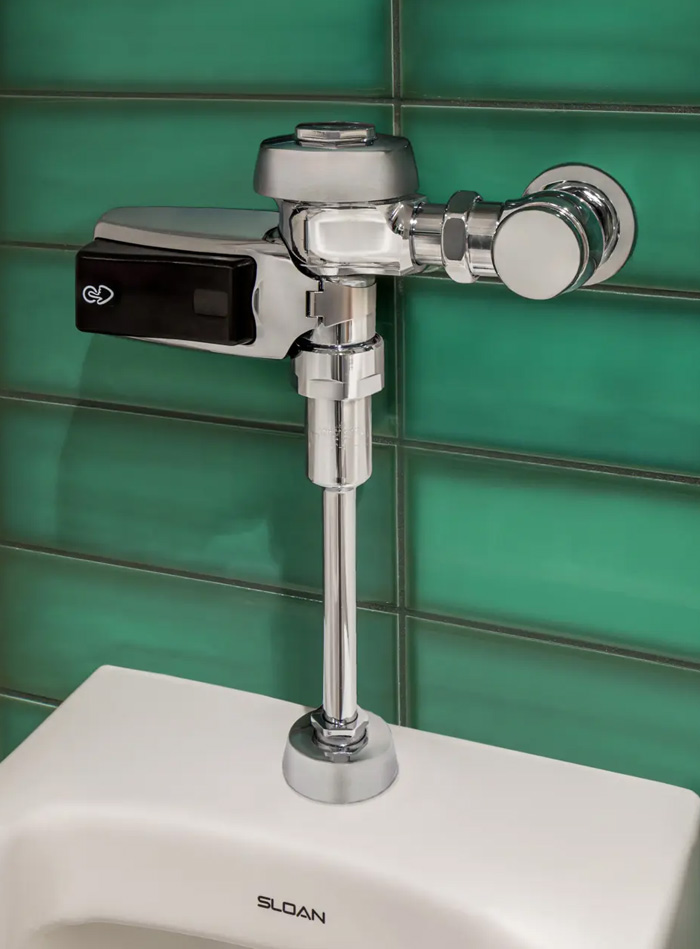 chrome Sloan automatic flush valve installed above a white toilet, set against a wall clad in glossy green rectangular tiles. The reflective metal finish contrasts with the vibrant background, conveying a sense of modernity, precision, and cleanliness.