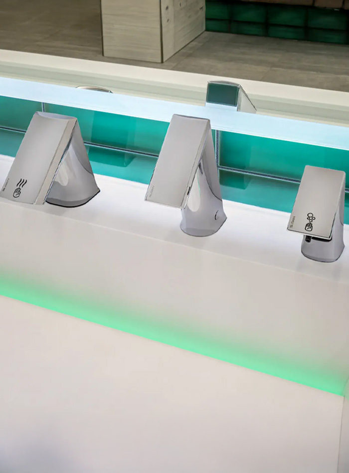 close-up of three Sloan automatic chrome faucets mounted on a white surface with integrated green LED lighting. The angular design of the fixtures and the reflection of the green-tiled background enhance the modern, high-tech aesthetic.