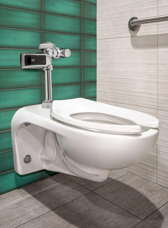 wall-mounted white porcelain toilet with a chrome Sloan automatic flush valve, set against a combination of glossy green tiles and light beige textured wall. The gray tile floor and stainless-steel grab bar complete a modern, functional, and visually balanced environment.