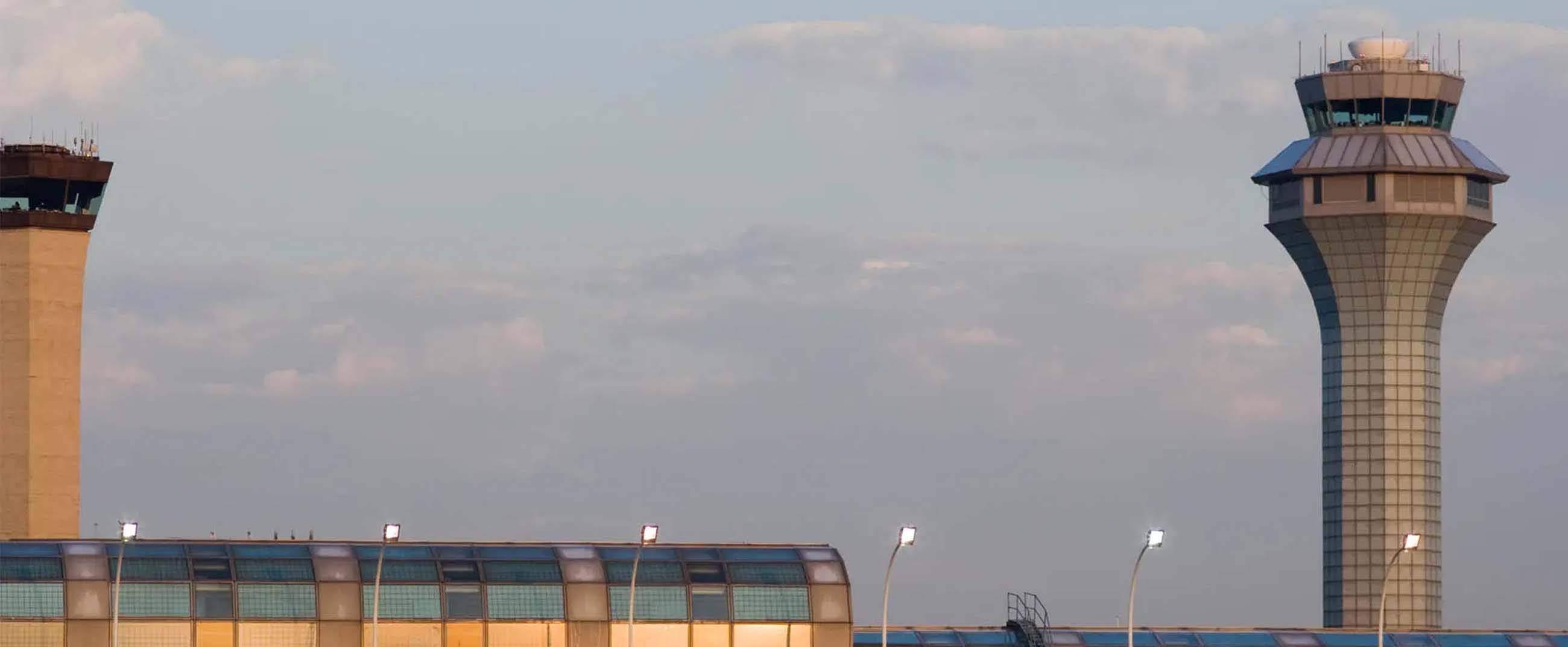 modern airport terminal at sunset, with two control towers visible in the background. The main structure features a curved design with reflective glass that captures the golden tones of the setting sun. The walkways and windows shimmer with warm light, while the towers—one taller and slimmer, the other more robust—stand out against the sky, symbolizing the airport’s operational core.