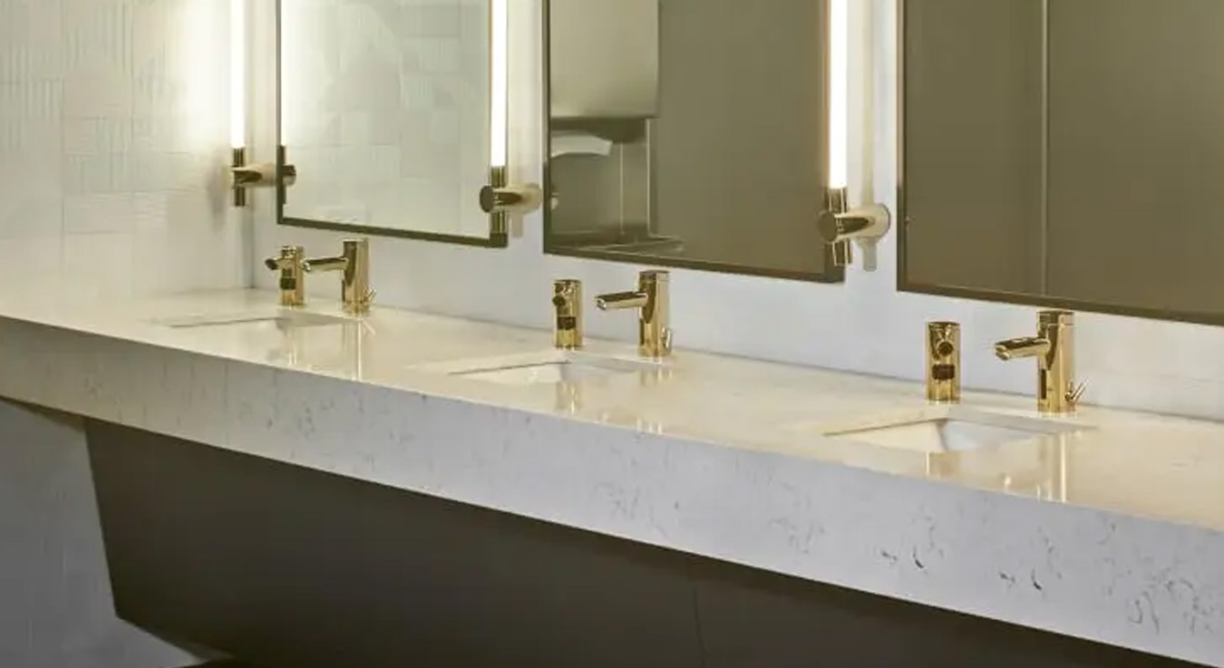 contemporary wash station with Sloan automatic faucets and soap dispensers in a gold finish, installed on a light marble countertop that enhances spaciousness and sophistication. The design blends understated luxury with intelligent functionality, featuring touch-free sensors that promote hygiene and water efficiency.