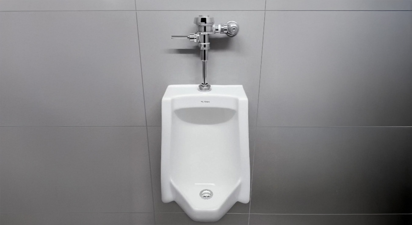a urinal with a Sloan manual flushometer, installed in a modern restroom with smooth gray wall finishes. Its minimalist and functional design highlights the durability of the materials and the efficiency of the hydraulic system, making it ideal for commercial or high-traffic environments.