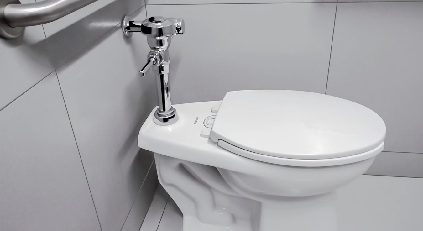 a floor-mounted toilet with a Sloan manual flushometer, installed in a modern accessible restroom. The smooth wall panels and stainless steel grab bars provide a clean, functional, and safe appearance, ideal for commercial spaces that prioritize accessibility and durability.