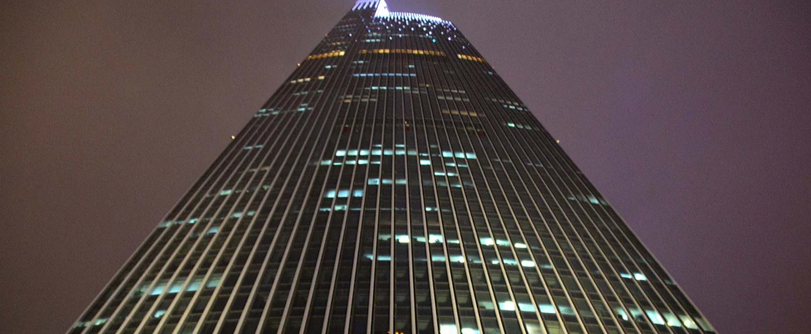 an impressive skyscraper captured from its base, highlighting its modern design with vertical lines that emphasize its height. The illuminated windows contrast against the night sky, creating an urban and elegant atmosphere.