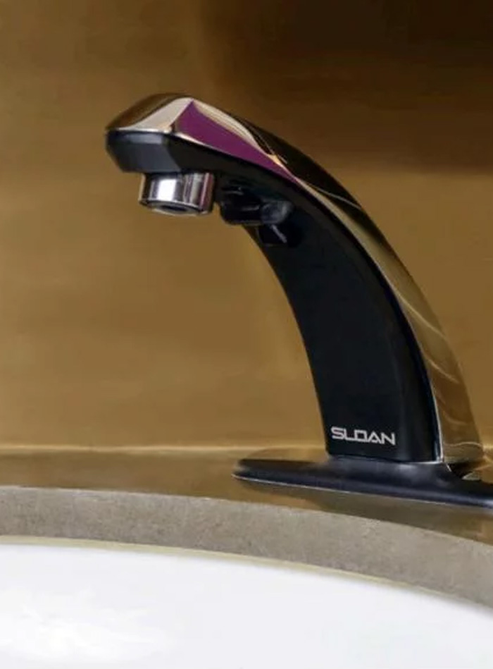 an elegant black Sloan automatic faucet with chrome details, installed on a smooth, modern countertop. The golden background enhances its sophisticated design and highlights the contrast between contemporary technology and luxurious finishes.