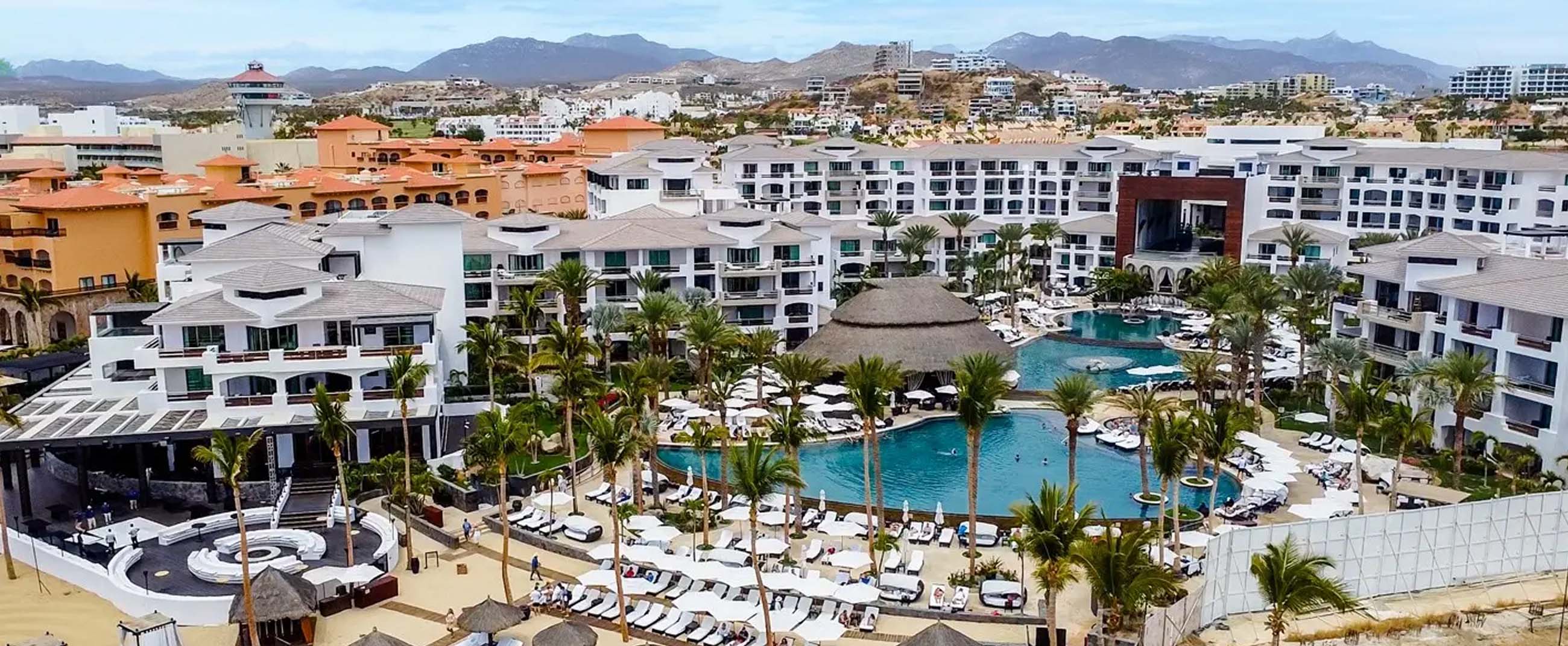 panoramic view of the Cabo Azul Resort Hotel, a luxury beachfront complex located in San José del Cabo, Mexico. The resort features contemporary architecture with tropical accents, expansive pools surrounded by palm trees and sun loungers, and a distinctive central palapa roof that serves as the visual centerpiece of the property.