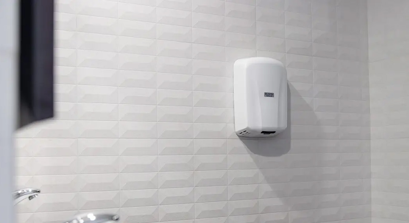 The image shows a Sloan XLERATOR® hand dryer mounted on a white geometric-tile wall at the Cabo Azul Resort Hotel. Its compact, sleek design blends seamlessly into the bathroom’s modern aesthetic, providing fast drying, low energy consumption, and touch-free operation, contributing to a more hygienic and sustainable guest experience.