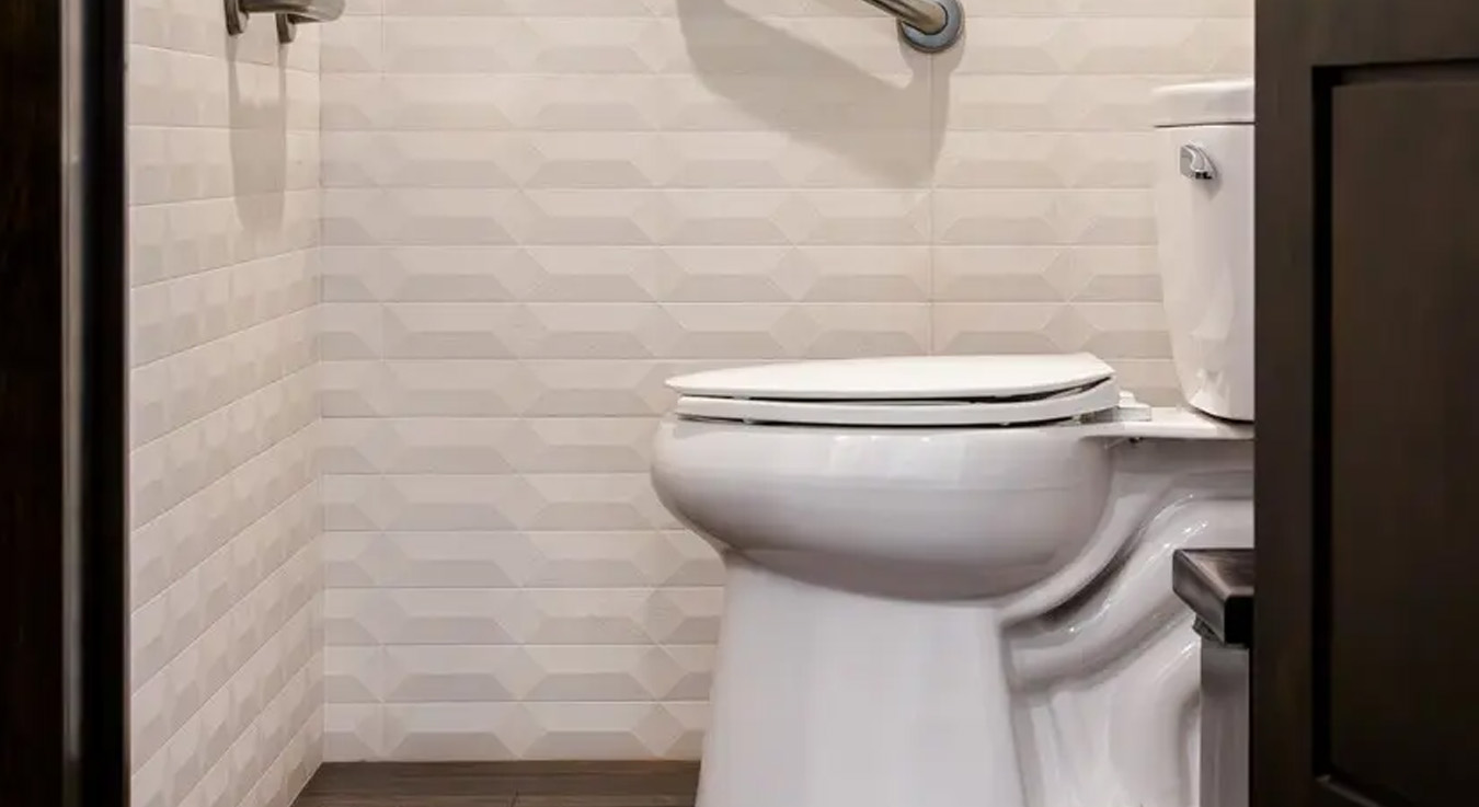 one-piece toilet with a clean, modern design, installed in a bathroom at the Cabo Azul Resort Hotel. The space features light-toned geometric wall tiles paired with a wood-like floor, creating a warm and elegant atmosphere. Additionally, the inclusion of stainless-steel grab bars enhances accessibility and user safety, reflecting the resort’s commitment to high-end hospitality standards.