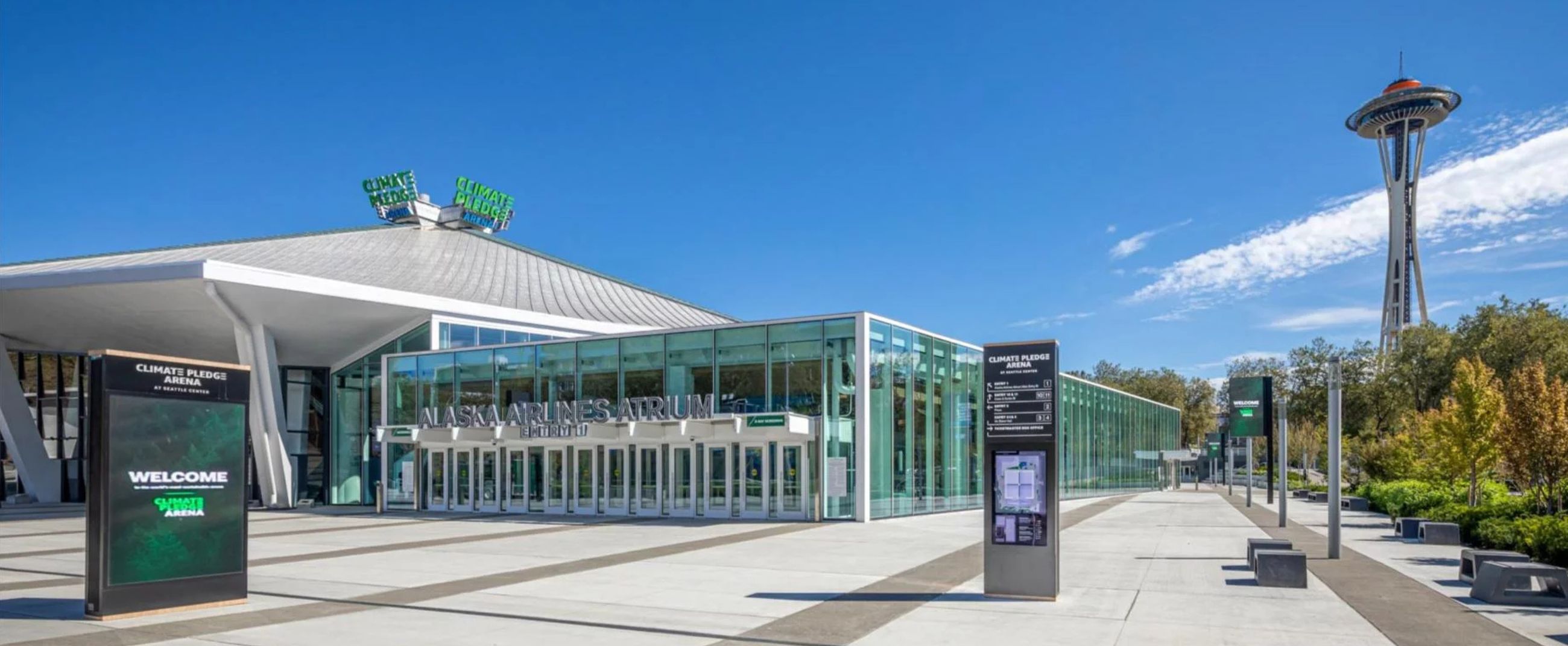 Main facade of Climate Pledge Arena in Seattle, featuring modern glass architecture with the iconic Space Needle in the background under a clear sky.