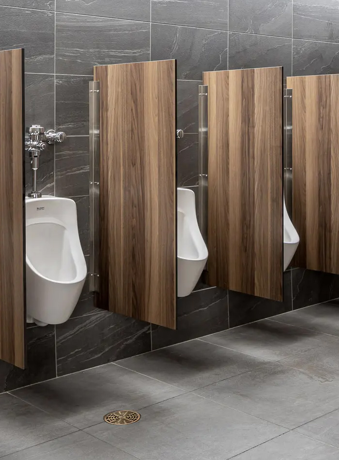 row of white Sloan urinals mounted on a dark gray tiled wall, separated by wooden privacy panels that add warmth and sophistication to the design. The combination of natural materials and modern finishes creates an elegant and functional space