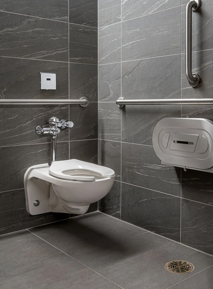 wall-mounted Sloan toilet installed in an accessible restroom, featuring stainless steel grab bars for safety and ease of use. The space is finished with large-format gray tiles, giving it a clean and modern look.