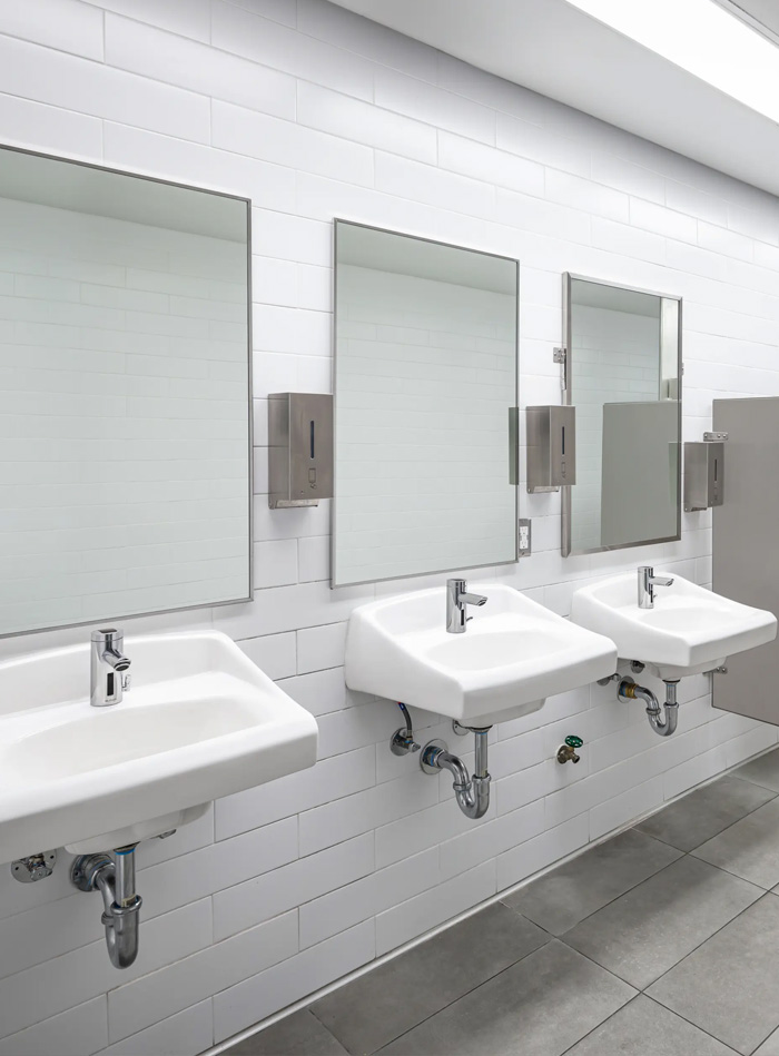 a modern, well-lit public restroom with a clean and functional design. It features two sinks with automatic faucets, soap and paper towel dispensers, and a urinal separated by privacy panels. The white walls and gray tiled floor enhance a minimalist and hygienic aesthetic.