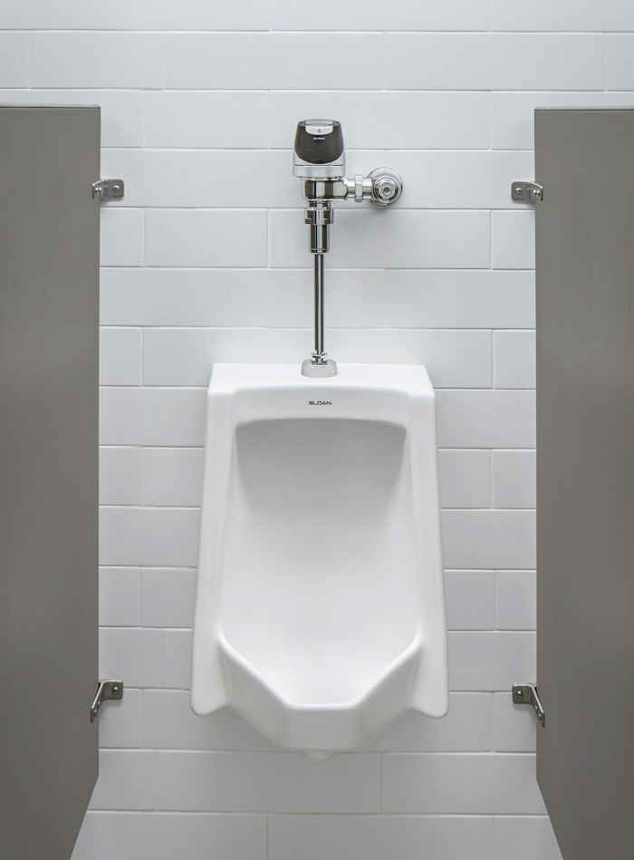a modern white urinal installed between two gray privacy panels. It features a chrome automatic sensor flush valve mounted on a wall covered with rectangular white tiles. The setting conveys cleanliness, functionality, and a contemporary style ideal for high-traffic public restrooms.