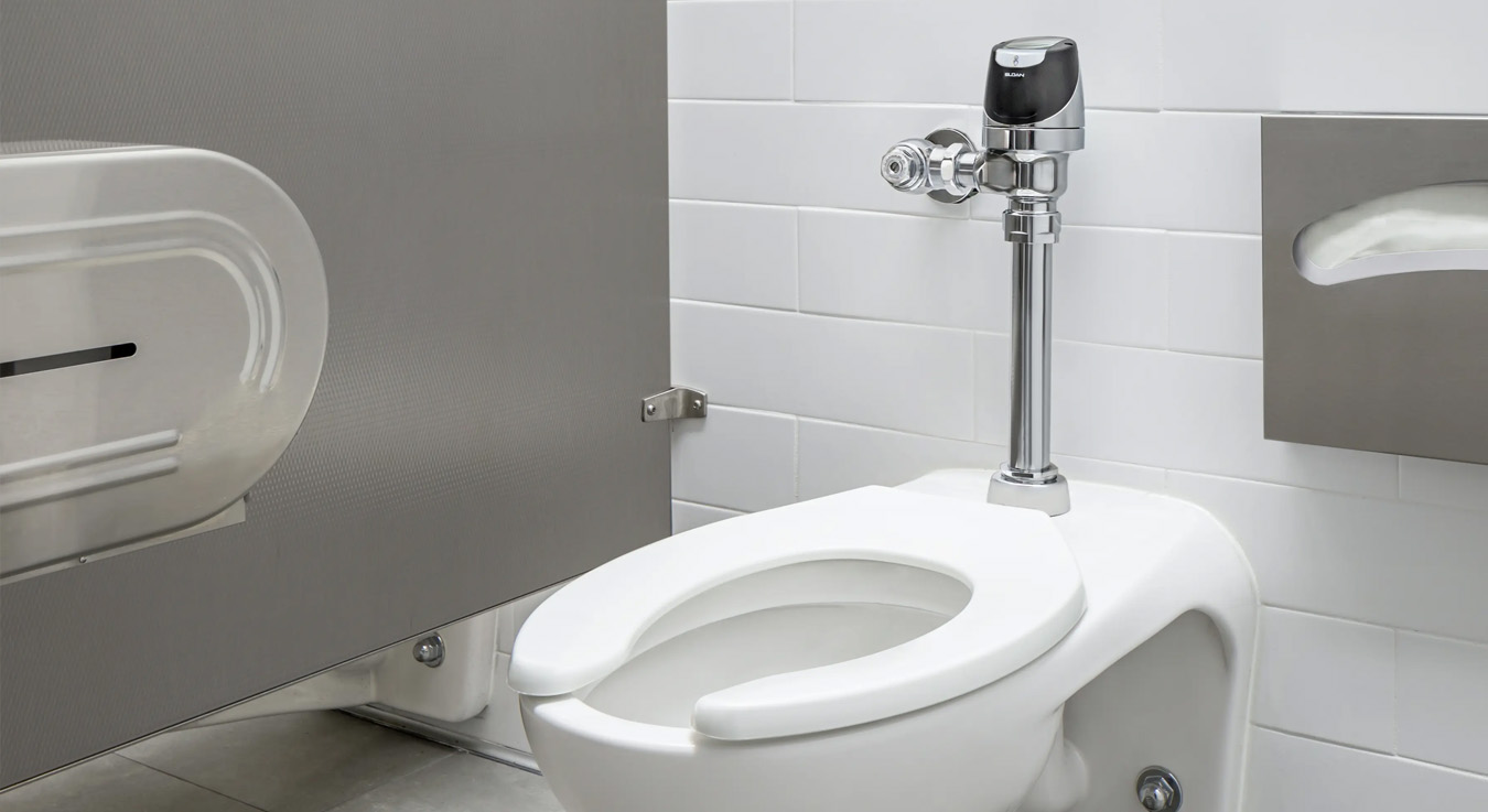 a wall-mounted toilet with a modern and sanitary design, featuring a chrome automatic sensor flush valve. It is installed in a clean, minimalist restroom with rectangular white tile walls, gray partitions, and gray floor tiles. A metal toilet paper dispenser is visible to the side, complementing the functional and contemporary aesthetic of the space.