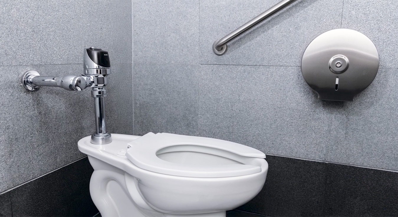 modern toilet with an automatic chrome flush valve and stainless steel accessories. The gray-toned environment conveys a sense of cleanliness and functionality, ideal for contemporary public spaces.