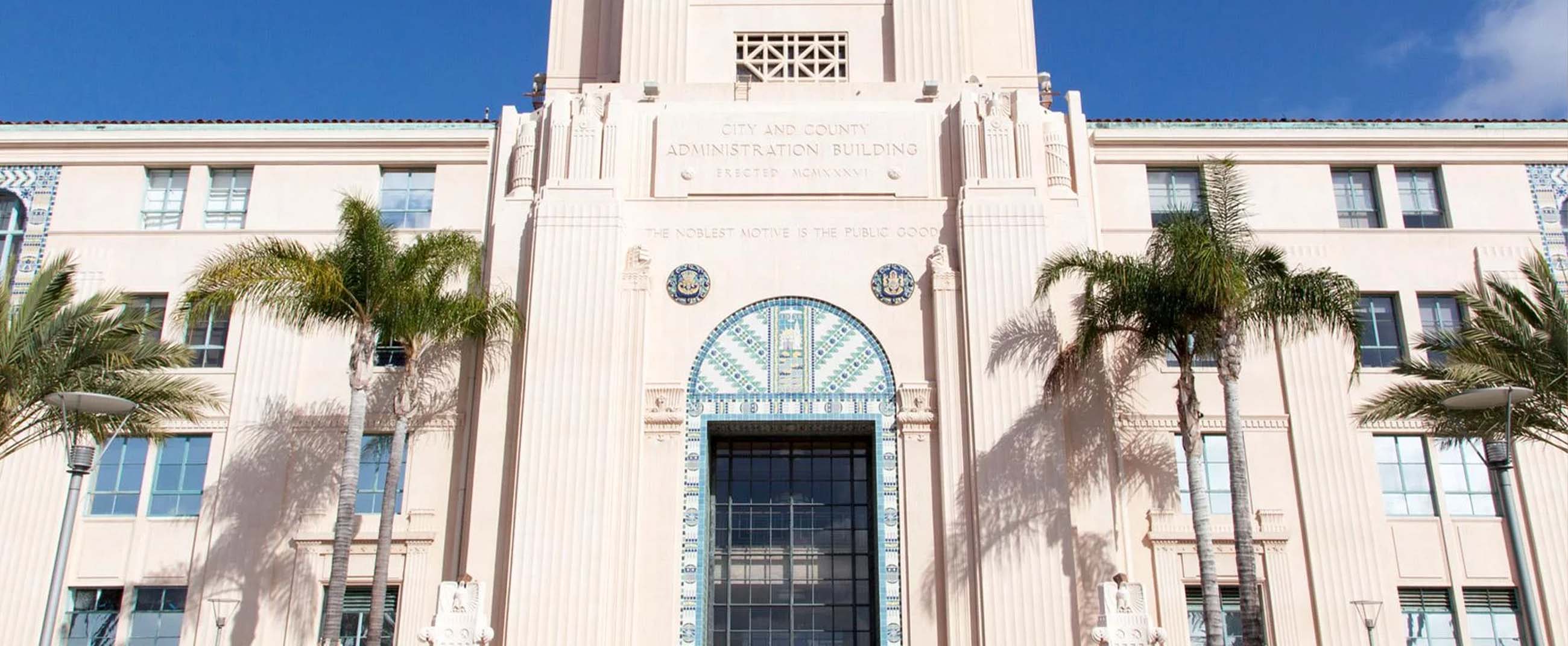 the City and County Administration Building in San Diego, California, an iconic Art Deco structure inaugurated in 1938. Its design features elegant vertical lines, blue ceramic details, and a grand main entrance symbolizing civic authority and public service. The façade bears the inscription “The Noblest Motive is the Public Good,” reflecting its role as an administrative center devoted to serving the community.