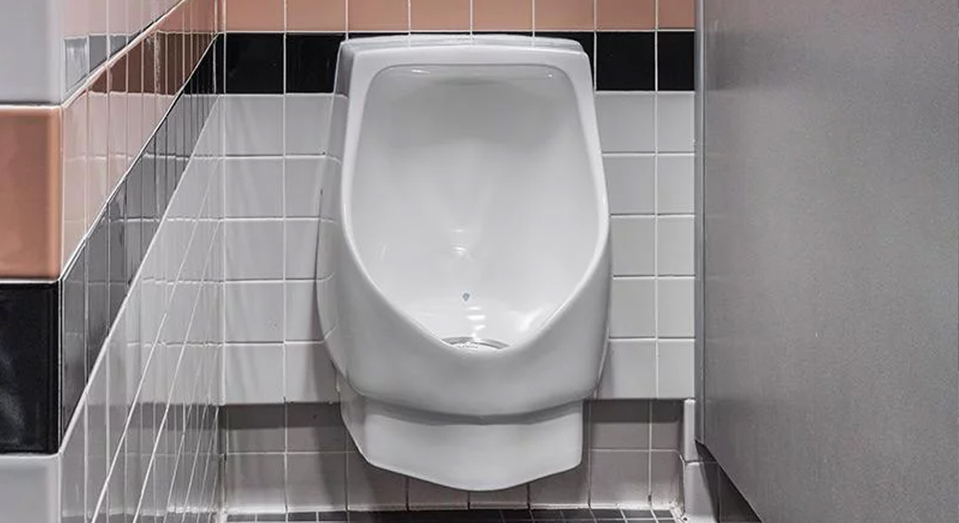 modern urinal installed in a public restroom featuring neutral-toned ceramic finishes with black and terracotta accents. Its recessed design maximizes space and simplifies cleaning, while the Sloan system ensures efficient, durable, and water-saving performance.