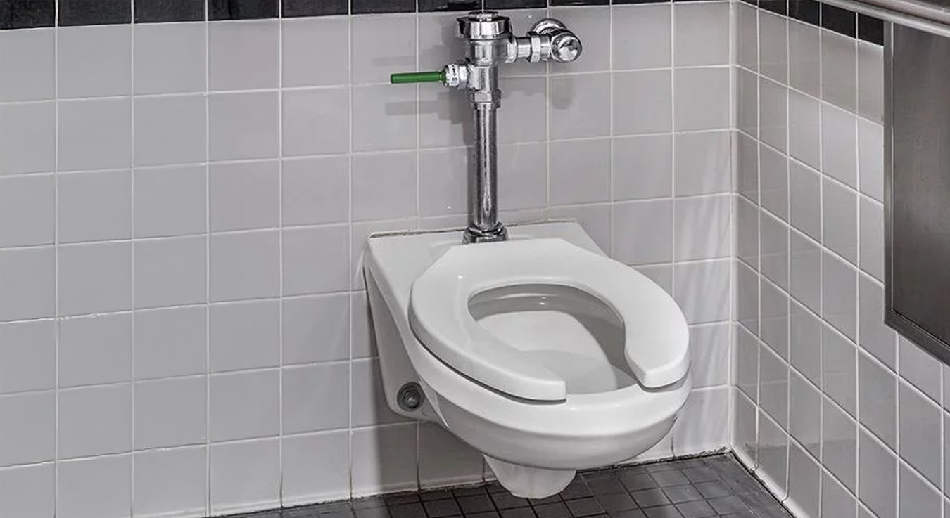 wall-mounted toilet with a manual flush valve, installed in a public restroom featuring neutral-toned ceramic finishes. The design includes stainless steel grab bars that meet accessibility standards, ensuring user comfort and safety. The integration of the Sloan system provides durability, easy maintenance, and water-use efficiency.