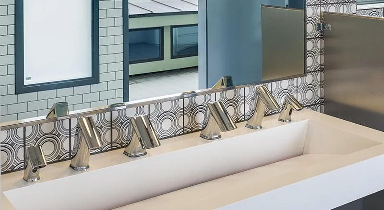 the washroom area at Hotel Erwin, featuring Sloan automatic faucets with a sleek, angular chrome design. The shared white solid-surface sink emphasizes minimalism, while the black-and-white circular-patterned tiles behind it introduce an artistic, retro flair that aligns with the hotel’s modern aesthetic. Overall, the design blends functionality, hygiene, and character, capturing the vibrant and contemporary spirit of Venice Beach.