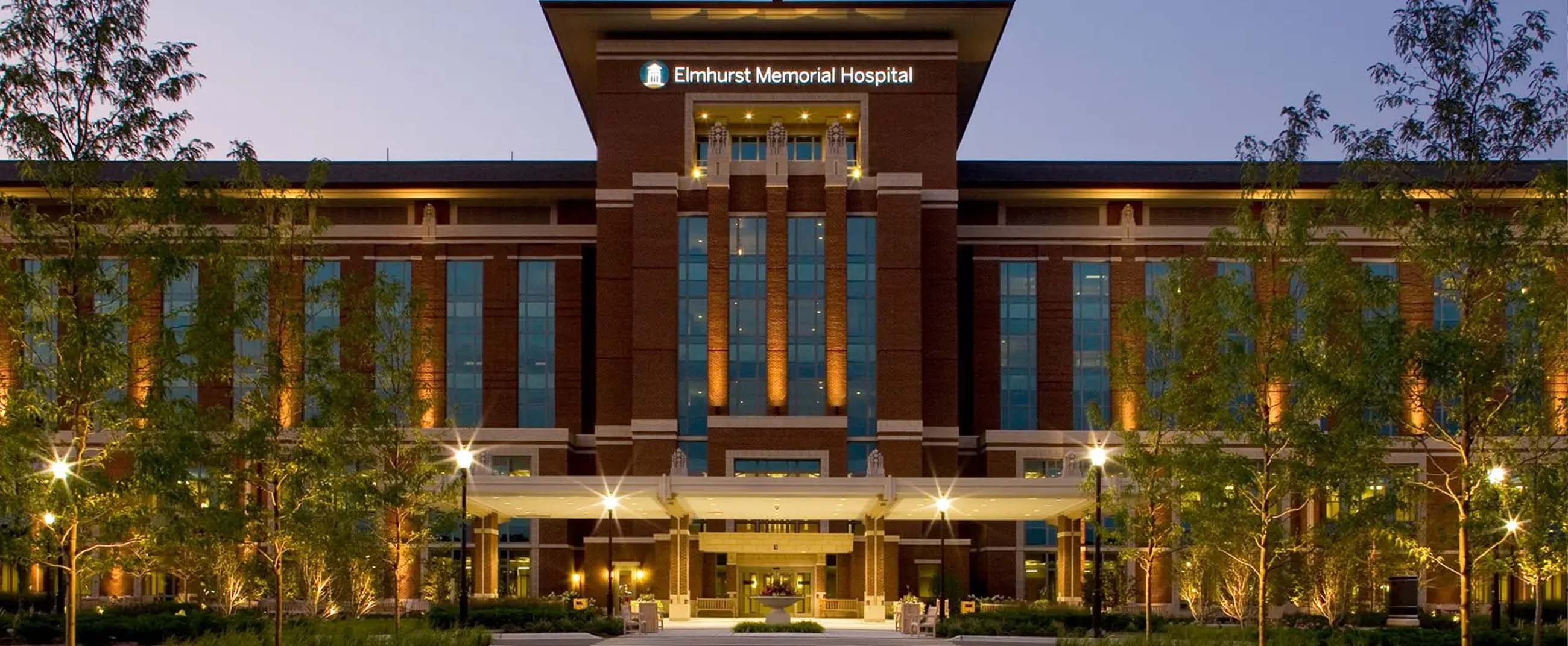 the main facade of Elmhurst Memorial Hospital, a modern brick building with tall vertical windows and warm lighting that highlights its architecture at dusk. The landscaped surroundings and symmetrical walkways create an atmosphere of calmness and professionalism.