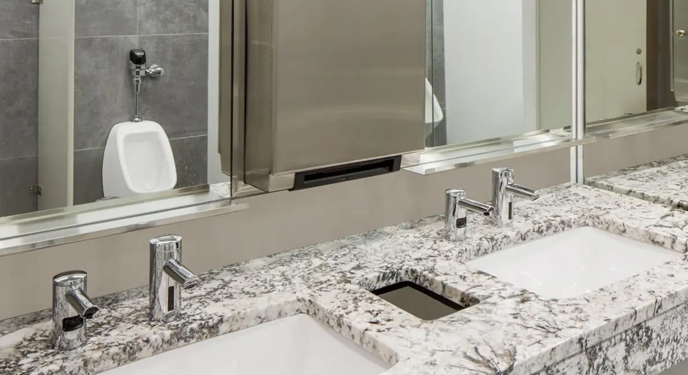 a high-end corporate restroom featuring Sloan automatic faucets and soap dispensers installed on a veined granite countertop, providing both elegance and durability. In the background, a sensor-activated urinal maintains visual and functional consistency.