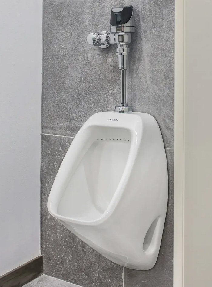a Sloan urinal equipped with an automatic sensor flushometer, providing touch-free hygiene and water efficiency. Its minimalist and functional design complements the gray wall finish, creating a modern and professional aesthetic.