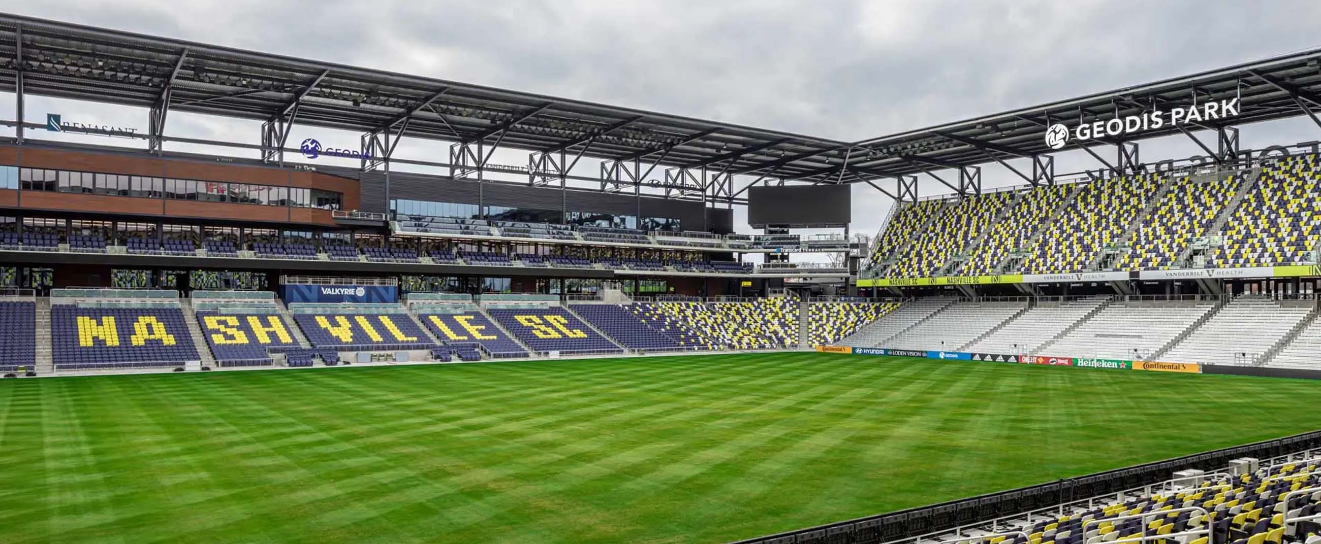 the interior of GEODIS Park in Nashville, home of Nashville SC. The stadium features a modern design with large covered stands, seats in shades of blue, yellow, and gray, and a meticulously maintained grass field. The architecture combines exposed steel and clean lines, reflecting a contemporary industrial style.