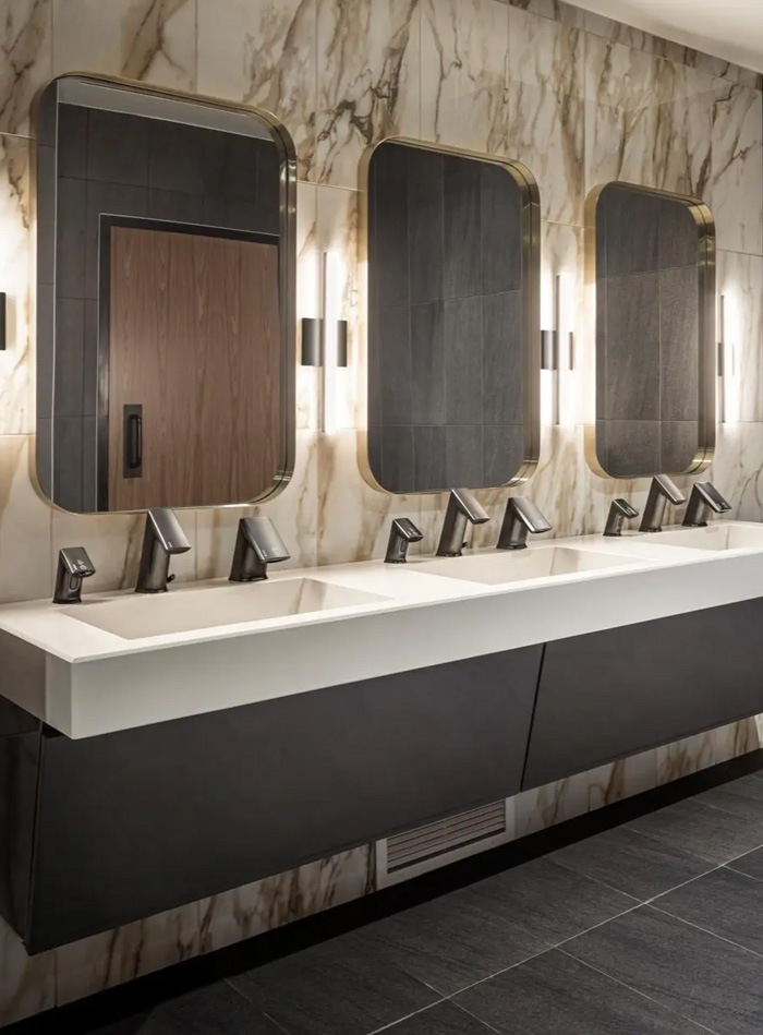 elegant contemporary restroom featuring a multi-station white solid-surface sink with dark metallic Sloan automatic faucets. The walls are clad in marble with golden and beige veining, while rounded mirrors with integrated vertical lighting add warmth and sophistication.
