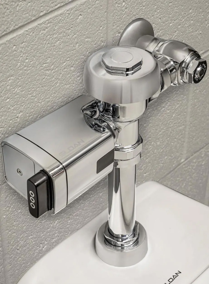 a chrome Sloan automatic flush valve mounted on a white toilet and attached to a gray block wall. The polished metal finish highlights the precision of the design and the quality of the materials, reflecting light and cleanliness.
