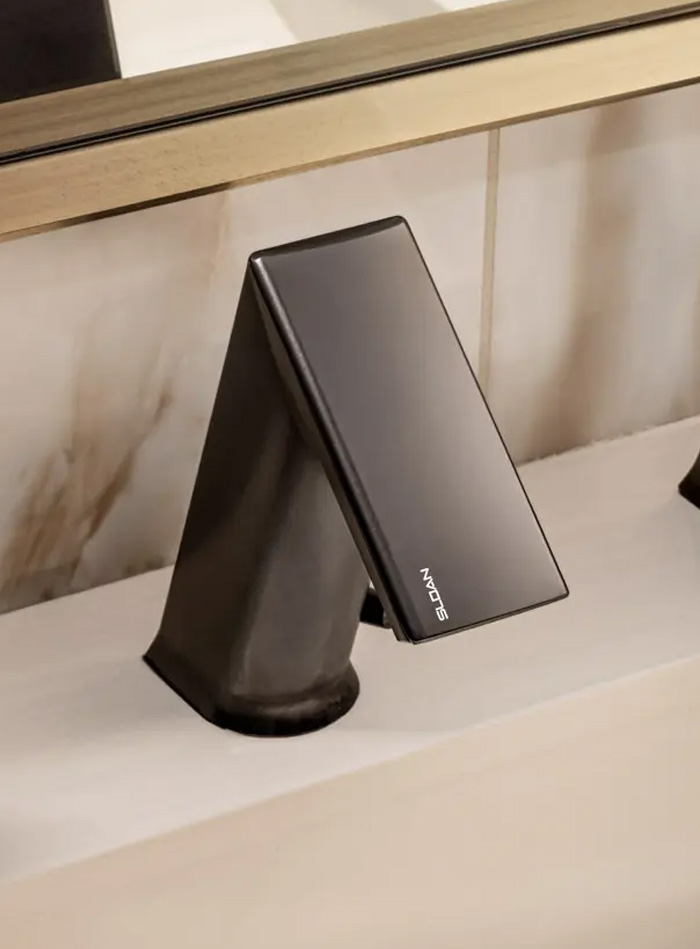 a Sloan automatic faucet in a dark metallic finish, mounted on a light surface beneath a gold-framed mirror. Its angular, minimalist design highlights precision and elegance,