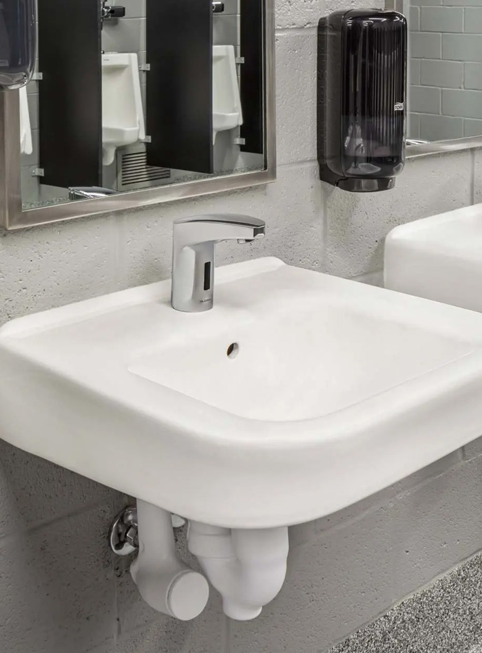 wall-mounted white porcelain sink with a chrome Sloan automatic faucet, installed against a painted gray block wall. A black wall-mounted soap dispenser is visible to the right, and urinals can be seen reflected in the metal-framed mirror.