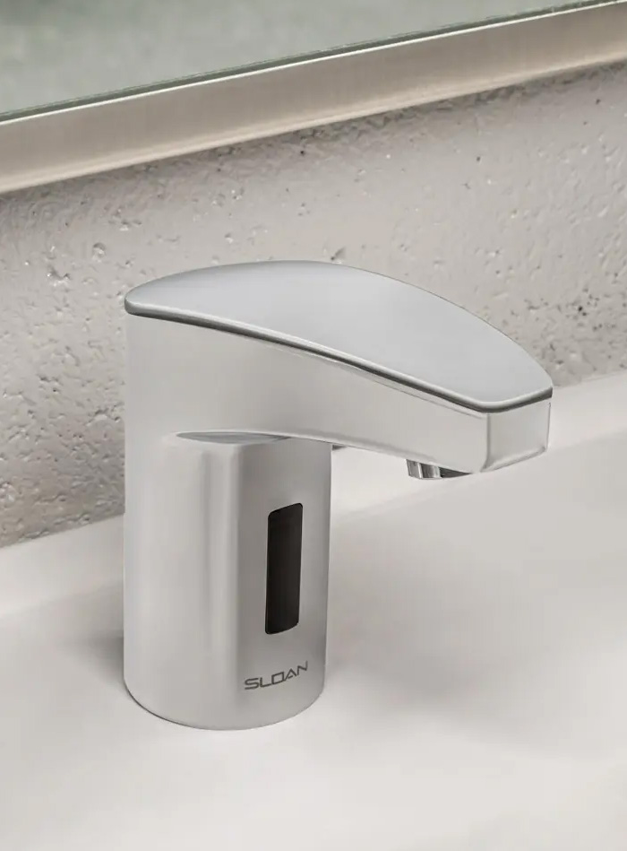 chrome Sloan automatic faucet with an integrated front sensor, mounted on a white surface and positioned in front of a metal-framed mirror. Its curved, compact design offers a modern and functional aesthetic, ideal for public spaces prioritizing hygiene, efficiency, and durability.