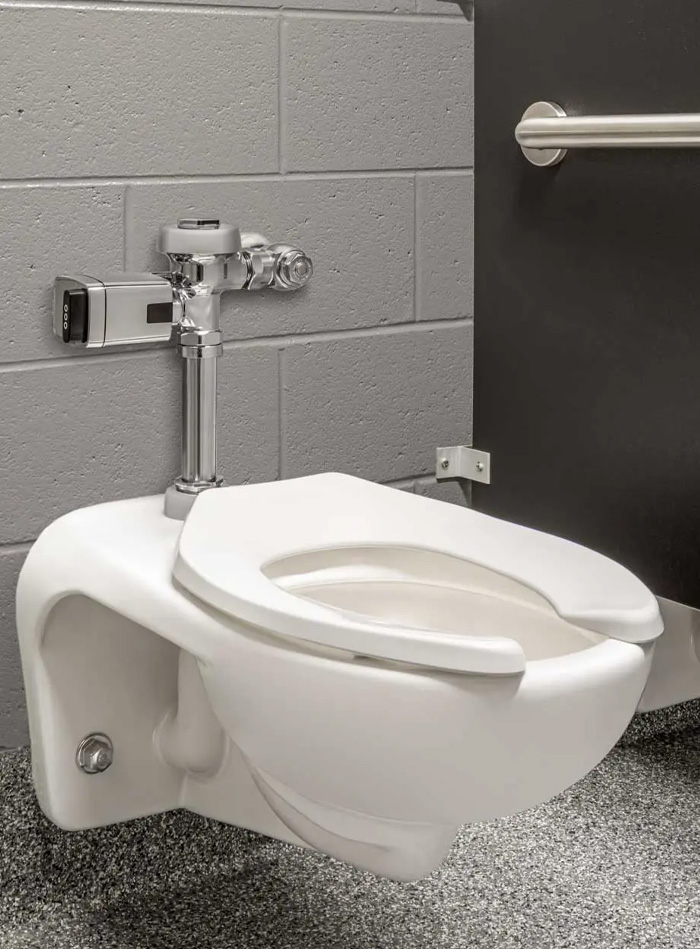 wall-mounted white porcelain toilet with a chrome Sloan automatic flush valve, installed against a painted gray block wall. A stainless-steel grab bar and dark stall door indicate an accessible restroom layout.