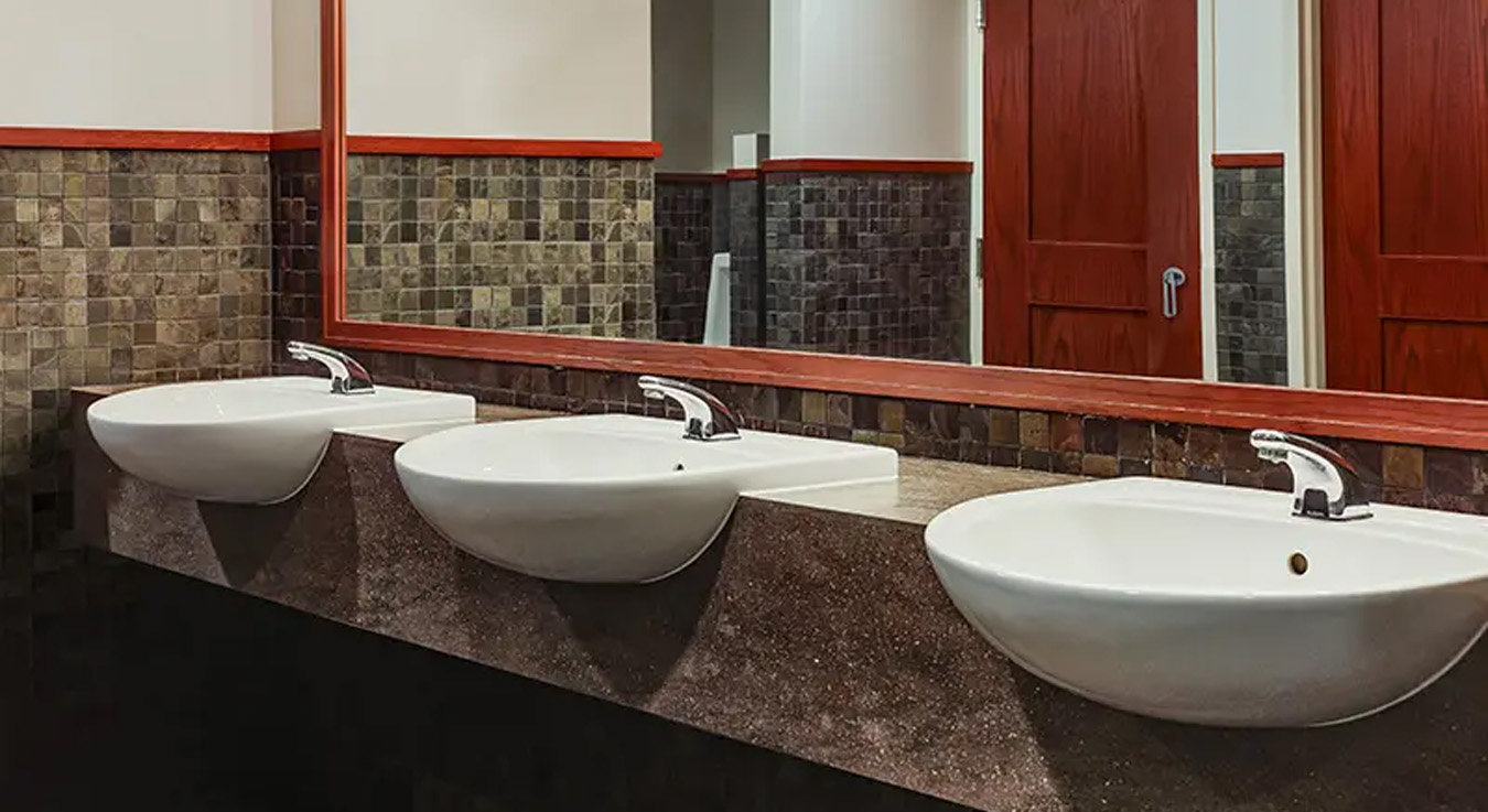 an elegant multiple-sink vanity with three white porcelain basins and automatic faucets, facing a large mirror with a wooden frame and warm lighting. The brown mosaic wall tiles and dark wood finishes create a sophisticated and welcoming atmosphere in the restroom.