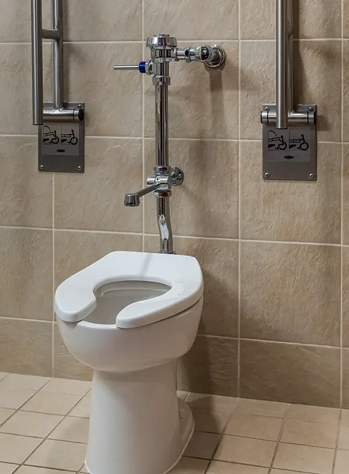 a public-use toilet designed for accessibility, featuring stainless steel side grab bars and a manual chrome flushometer. The setting, with beige tile walls, conveys cleanliness and functionality, ideal for institutional or commercial spaces.